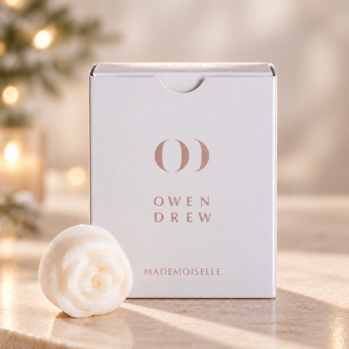 Owen Drew Mademoiselle wax melt box beside a white rose-shaped melt with softly lit Christmas greenery