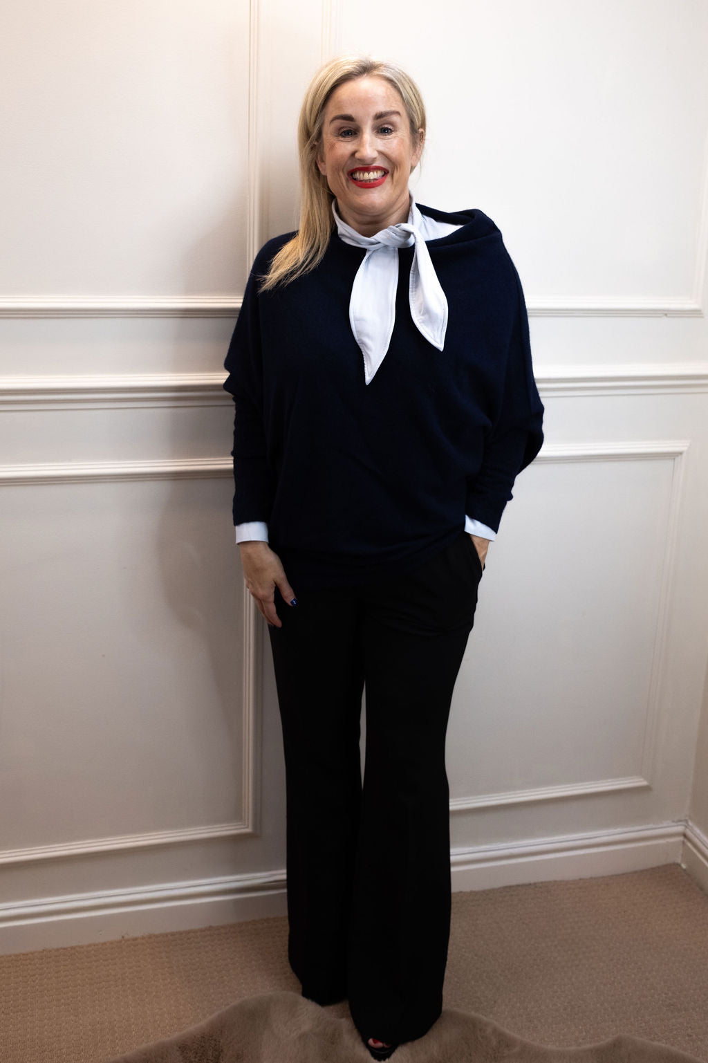 Woman wearing the light blue Sofia neck-tie shirt with a navy knit and black trousers, hands at her pockets.