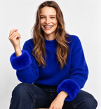 Seated model wearing royal blue faux fur wrist cuffs with a matching blue jumper and dark jeans.