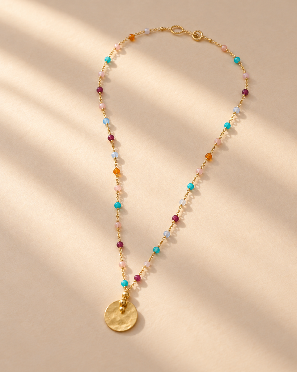 Sorbet necklace with multicoloured beads and round gold-tone pendant on a cream surface.