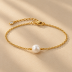 Gold-plated fine-chain bracelet with a single white pearl, shown on a warm cream surface.
