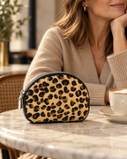 Bari leopard-print small purse on a marble cafe table beside a woman in a neutral knit, with black edging and silver zip visible.