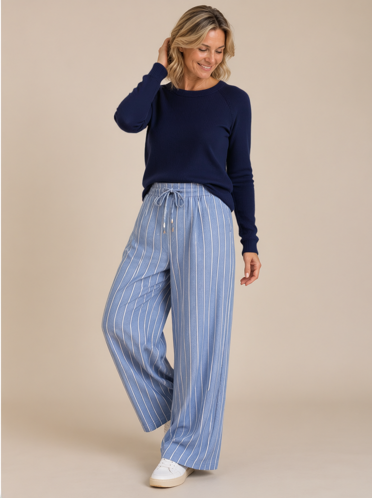 Woman over 50 modelling Bonnie cashmere blue and cream fine striped wide-leg trousers with a gathered drawstring waist, navy knit and white trainers against a warm neutral studio backdrop.
