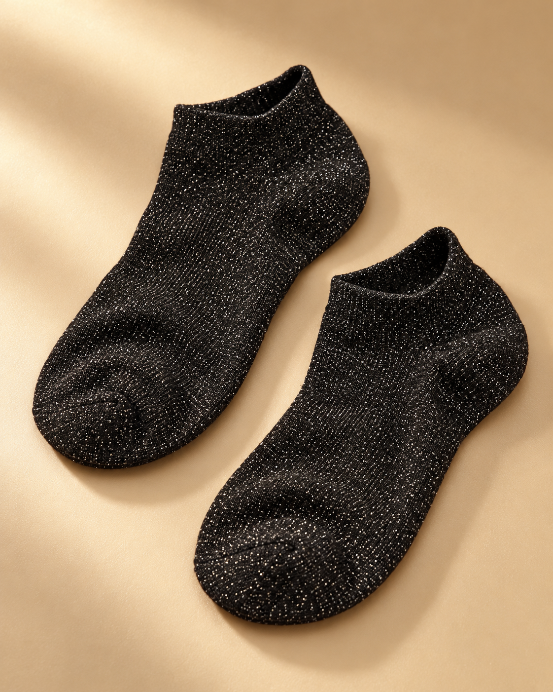 Pair of black glitter sneaker socks laid flat on a warm golden minimalist studio background