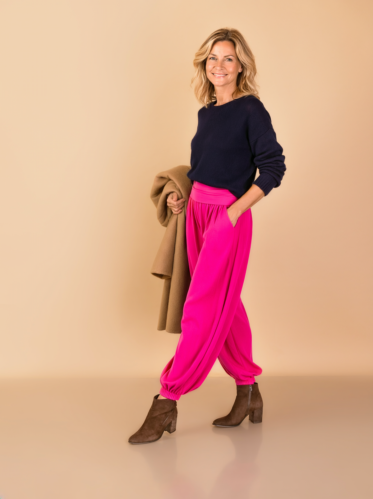 Woman over 50 modelling vivid fuchsia Tia harem trousers with a folded waistband and gathered ankle cuffs, paired with a navy jumper and brown ankle boots against a neutral studio background.
